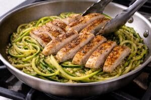 Sliced chicken being added back to a skillet with zucchini noodles and garlic, gently tossed with tongs and lightly coated in olive oil with slight steam visible