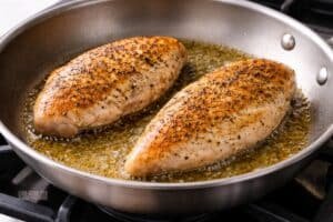Chicken breasts cooking in a skillet with olive oil, showing golden brown sear and small bubbles of oil around the edges on a clean stovetop