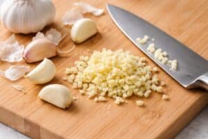 Garlic cloves being peeled and finely chopped on a cutting board with a chef’s knife, showing whole cloves, partially peeled garlic, and evenly minced pieces