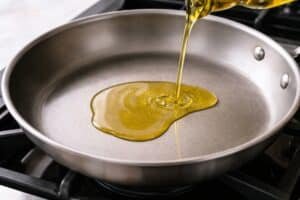 Olive oil being poured into a heated skillet on a stovetop, spreading and lightly shimmering across the pan surface in a clean kitchen setting
