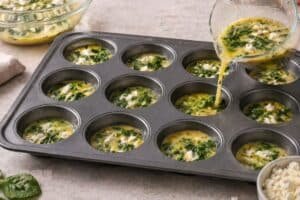 pouring egg mixture with spinach and feta into 12 cup muffin tin on kitchen countertop