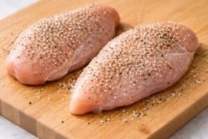 Raw chicken breasts on a cutting board seasoned with salt and black pepper, showing visible grains and surface texture in a clean kitchen setting