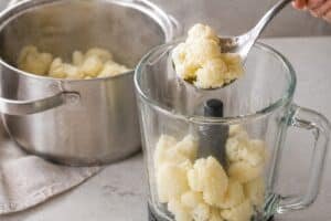 steamed cauliflower florets being transferred from a pot into a blender with a spoon, light steam visible in a minimal kitchen setting