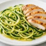 zucchini noodles with olive oil and sliced chicken on a white plate with garlic pieces visible