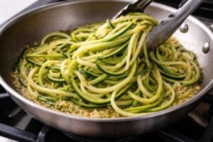 Zucchini noodles being added to a skillet with garlic and olive oil, lightly sautéing and gently stirred with tongs, showing glossy texture in a clean kitchen setting