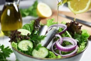 Drizzling olive oil and fresh lemon juice over sardine salad with cucumber, parsley, and leafy greens in a large bowl