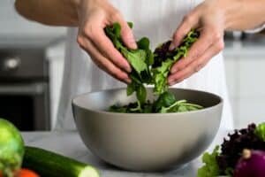 Fresh leafy greens being added into a large mixing bowl in a bright home kitchen with natural healthy ingredients nearby