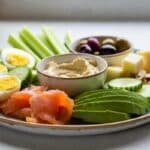Low-carb snack plate for steady energy featuring eggs, smoked salmon, avocado, cucumber, olives, cheese, and nuts arranged in a bright kitchen setting.