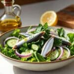 Sardine salad with olive oil and lemon served with leafy greens, cucumber, parsley, and red onion in a ceramic bowl