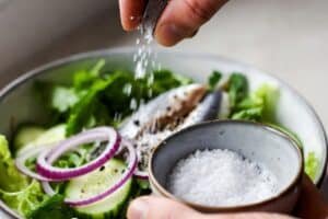 Seasoning sardine salad with sea salt and black pepper over leafy greens, cucumber, and red onion in a ceramic bowl